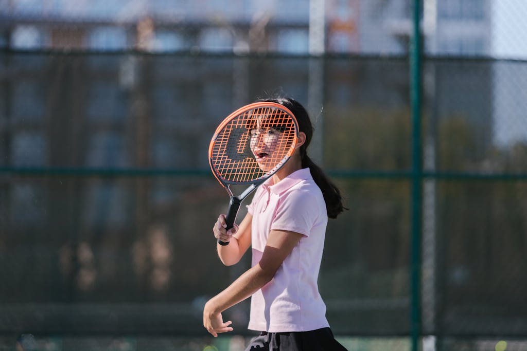 A girl in sportswear practices tennis on an outdoor court in sunny weather.